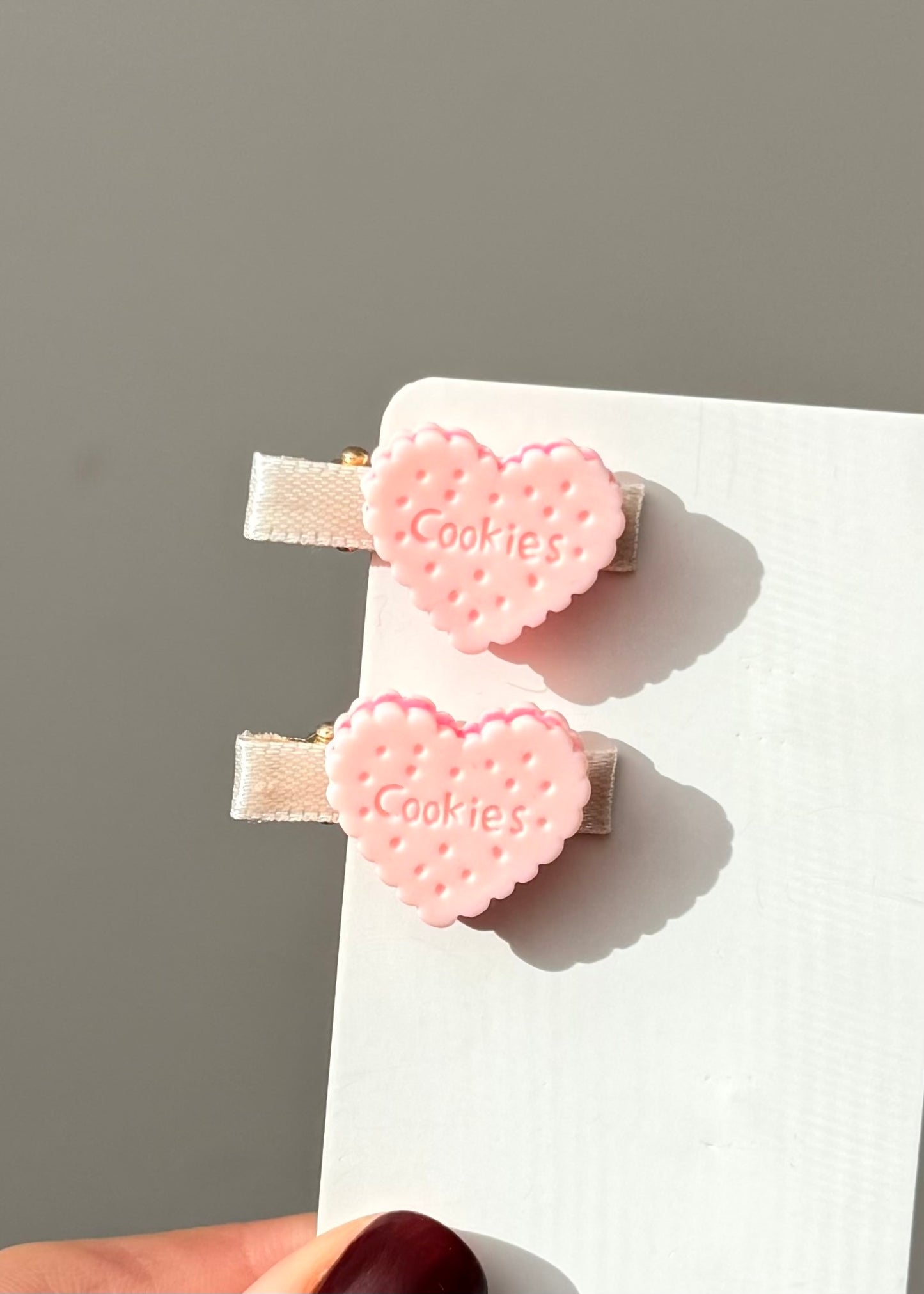 Two pink heart-shaped hair clips with 'Cookies' text on a white card against a gray background.