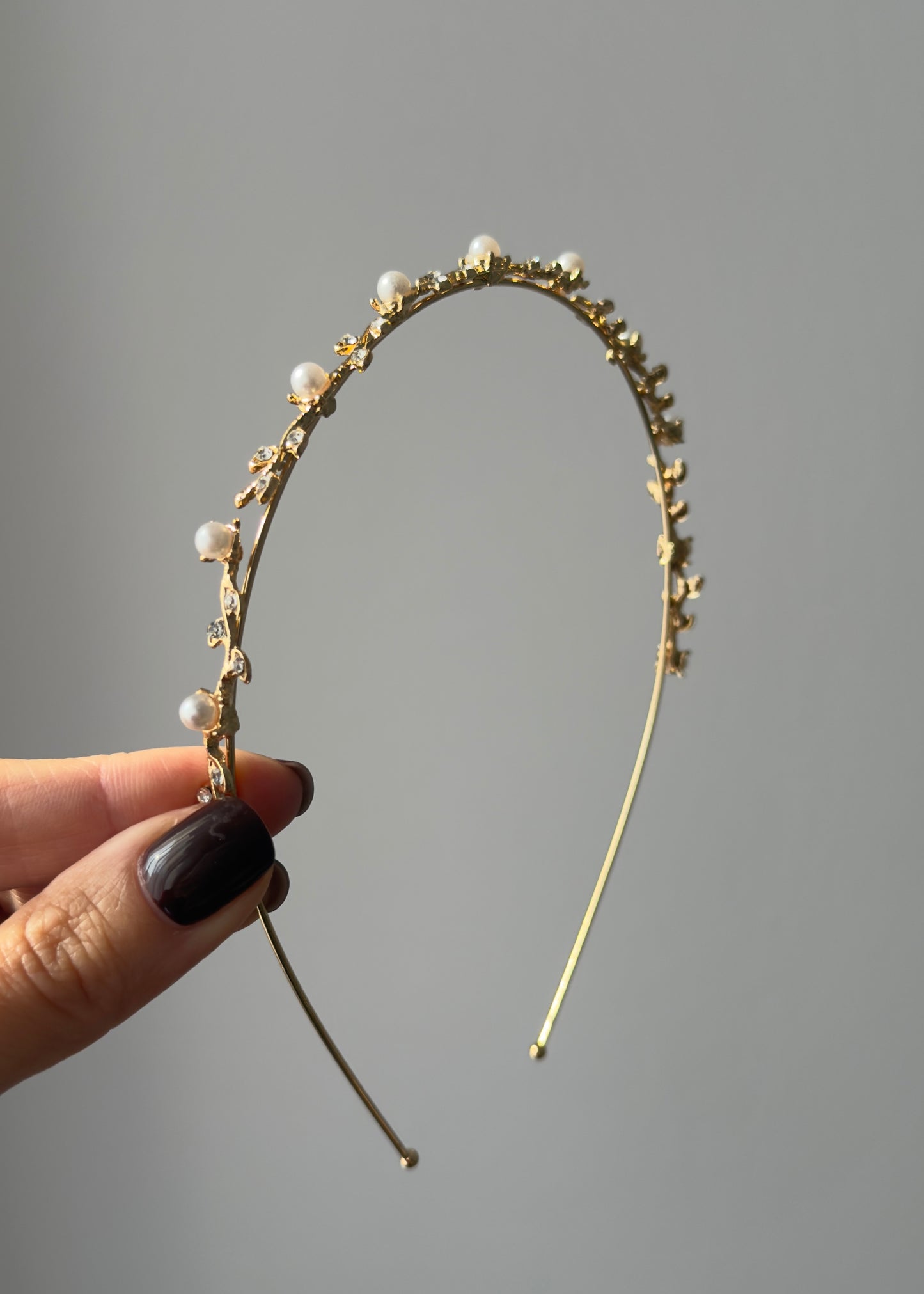 Gold headband with pearls held by a hand against a gray background